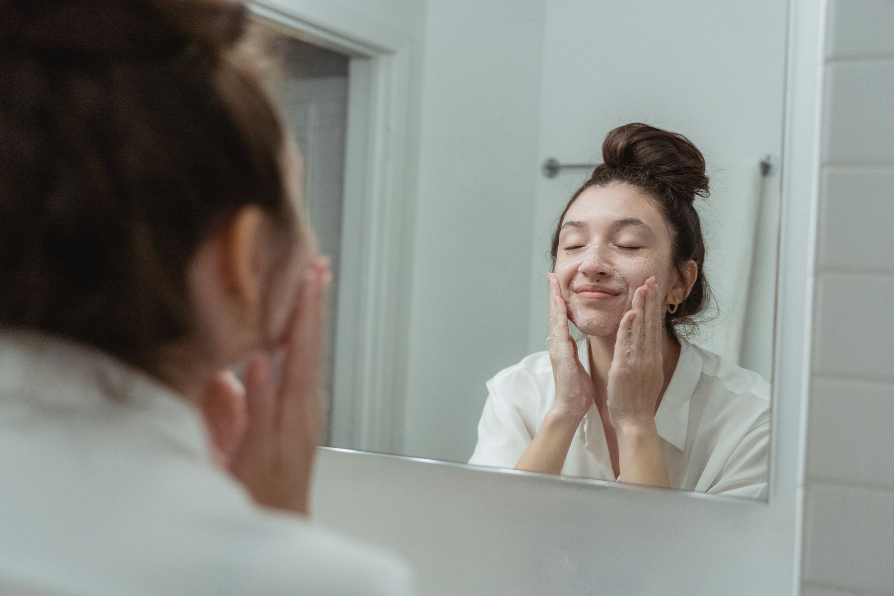 Woman applying cleanser to her cheeks with her fingers in a bathroom mirror.